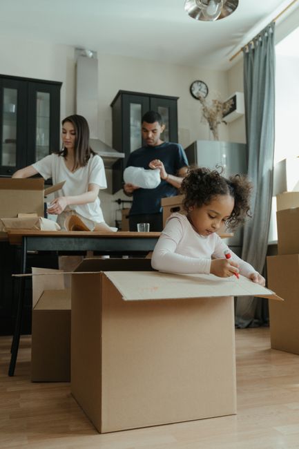 A young girl with curly hair sitting at a table inside a home, drawing on a piece of cardboard with a red marker, surrounded by open cardboard boxes and packing materials, as part of a home relocation