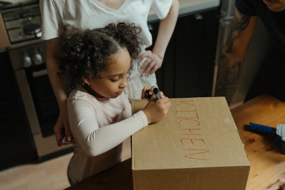 A young girl with curly hair sitting at a table inside a home, drawing on a piece of cardboard with a red marker, surrounded by open cardboard boxes and packing materials, as part of a home relocation