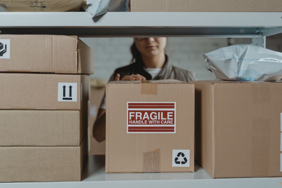 A person wearing a dark blue shirt is packing fragile glass bottles into a cardboard box lined with white packing peanuts, inside a well-lit room with wooden flooring. The individual is carefully plac